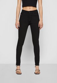 Nő magas derekú fekete skinny farmernadrágban, fekete crop topban és fekete tűsarkú, szegecses szandálban, egy sima világos háttér előtt áll.