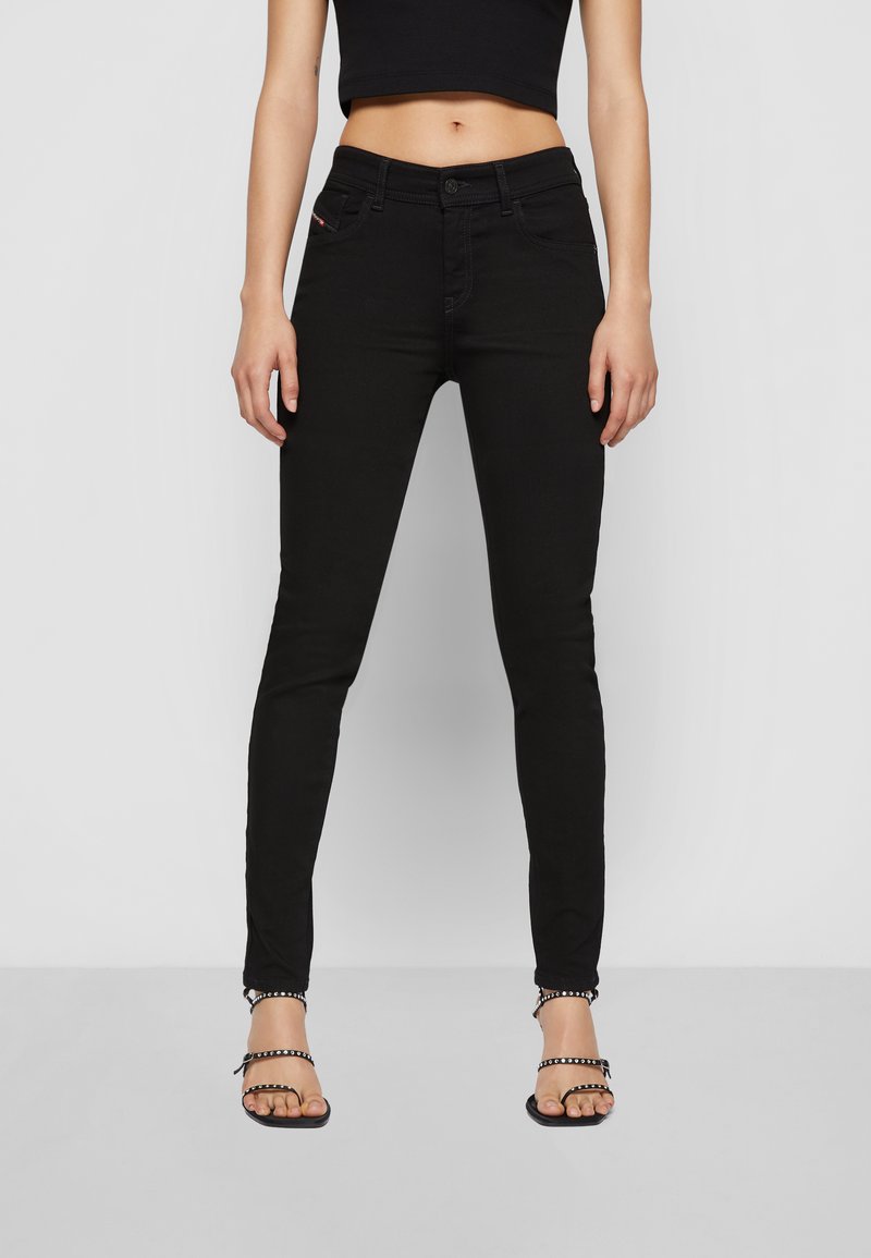 Nő magas derekú fekete skinny farmernadrágban, fekete crop topban és fekete tűsarkú, szegecses szandálban, egy sima világos háttér előtt áll.