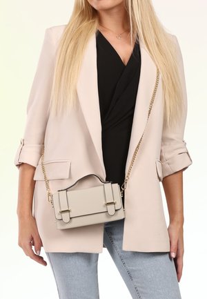 Femme portant un blazer beige clair, un top noir, un jean bleu et portant un petit sac beige avec une bandoulière chaîne dorée en travers du corps.