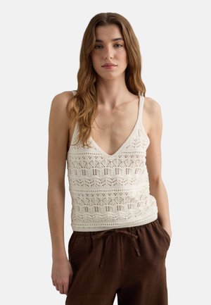 Femme aux longs cheveux châtain clair portant un haut blanc en crochet sans manches et un pantalon marron foncé à cordon, debout avec une main dans la poche.