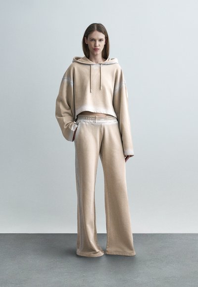 Γυναίκα που στέκεται φορώντας μπεζ φαρδιά crop hoodie και αντίστοιχο παντελόνι φαρδύ, μπροστά σε απλό ανοιχτό γκρι φόντο.