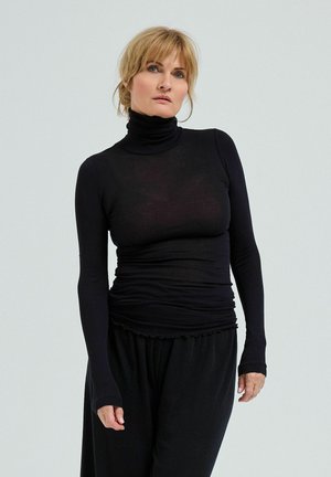 Ženska s svetlimi lasmi, oblečena v oprijet črn pulover z visokim ovratnikom in široke črne hlače, stoji pred enakomerno svetlo sivo ozadje.