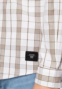 Chemise à carreaux blancs avec un motif en plaid marron, comportant une étiquette en tissu noir avec le texte "S. OLIVER ELEVATED CASUAL" sur l'ourlet.