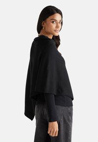 Černý pončo s volným, splývavým designem, vyrobené z měkké tkaniny. Má široké rukávy a je vrstveno přes černý dlouhý rukáv a šedé kalhoty.