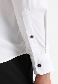 Cuff di una camicia bianca a maniche lunghe con un bottone nero e un bottone di accento rosso, realizzato in tessuto liscio e con una vestibilità su misura.
