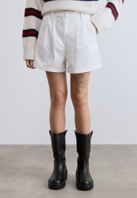 Λευκά σορτς με ψηλή μέση και πιέτες μπροστά, συνδυασμένα με μαύρες αδιάβροχες μπότες μέχρι το γόνατο και ένα ζεστό πλεκτό πουλόβερ με κόκκινες ρίγες.