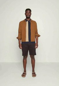 Jaqueta castanha sobre um polo às riscas bege e azul-marinho, combinado com calções pretos e sandálias castanhas. O conjunto apresenta um corte casual e descontraído.