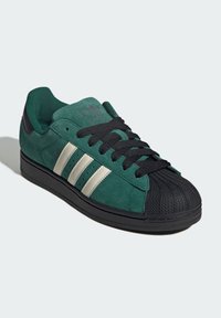 Groene suède sneaker met een zwarte rubberen neuskap en buitenzool. Heeft drie witte strepen, zwarte veters en een gewatteerde kraag.
