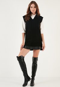 Black knitted sleeveless dress with a v-neck and ribbed texture, layered over a white collared shirt. Features a pleated skirt and paired with black thigh-high boots.