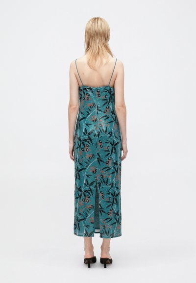 Žena stoji okrenuta leđima, obučena u tirkiznu cvjetnu slip haljinu s tankim trakama i crnim potpeticama, nasuprot jednostavnoj bijeloj pozadini.