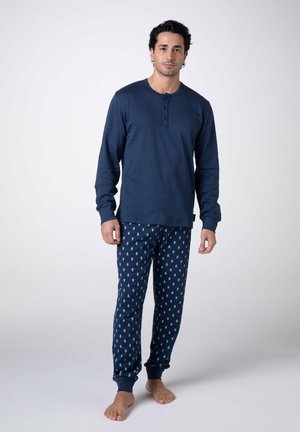 Uomo in piedi a piedi nudi che indossa una maglia henley a maniche lunghe blu navy e pantaloni da pigiama blu navy con motivo paisley azzurro.
