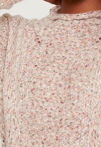 Vaaleanbeige neulepaita, jossa on pilkullisia tekstuureja vaaleanpunaisessa, oranssissa ja ruskeassa. Pyöreä kaula-aukko hienovaraisella resorireunuksella.