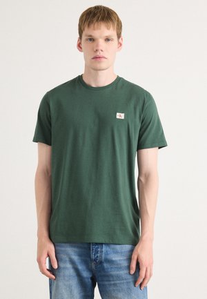 Camiseta de algodón verde con cuello redondo, mangas cortas y un pequeño parche de logo en el lado izquierdo del pecho. Textura suave, corte clásico.