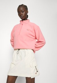 Sudadera de forro polar rosa con cuello alto y media cremallera, combinada con una falda de un beige claro que presenta bolsillos y detalles de cuerda.