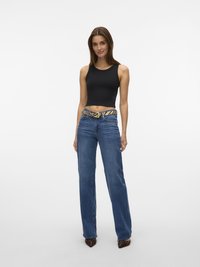 Svart crop top, blå vide jeans og et stripete belte i brunt og svart. Modell iført brune hæler med spiss tå. Enkelt bakgrunn.