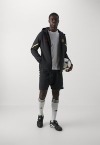 Schwarze Sportjacke mit gelben Akzenten, graues T-Shirt, schwarze Shorts und weiße Socken mit schwarzen Streifen. Hält einen Fußball.