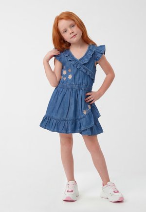 Giovane ragazza dai capelli rossi che indossa un vestito blu con balze e ricami floreali e sneakers bianche, posando con una mano sul fianco e l'altra che le tocca i capelli.