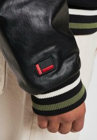 Sort læder bomberjakke med ribbede sort, hvid og olivengrønne manchetter; har et firkantet logo-patch i rød og sort på ærmet.