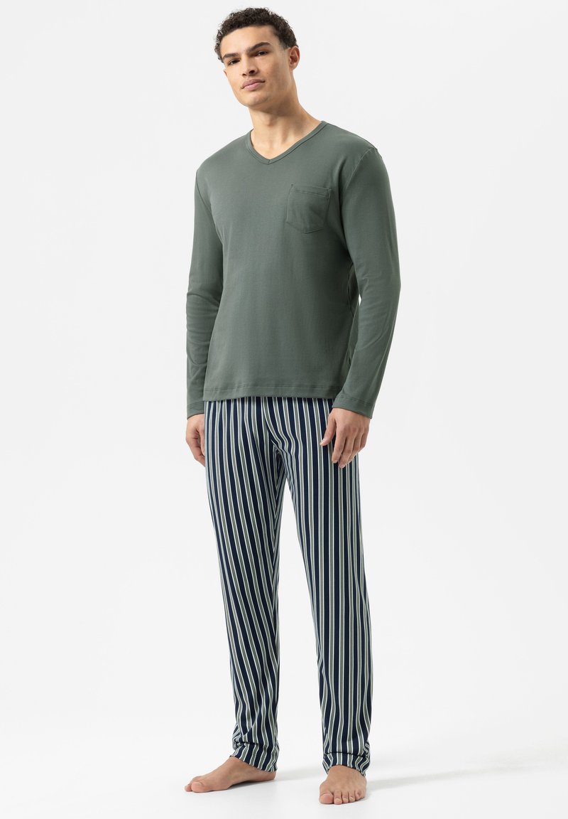 Man staat blootsvoets en draagt een groene lange mouw V-hals shirt en een donkerblauwe en witte verticaal gestreepte pyjamabroek.