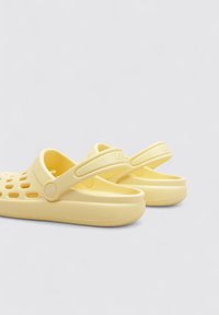 Sabots en plastique jaune avec un dos fermé, un dessus perforé pour la respirabilité et une semelle lisse et plate. Marque visible sur la sangle.