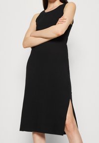 Robe noire sans manches en coton, avec un col rond et des fentes latérales pour faciliter les mouvements. Design ajusté avec un minimum de détails.