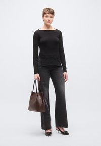 Fekete hosszú ujjú felső hajónyakú kivitelben, sötétszürke, szárában bővülő farmerral párosítva; a modell egy sötétbarna, vállpántos fonott táskát tart.