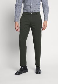 Pantalon vert foncé ajusté en tissu lisse, avec fermeture à bouton et poches latérales, associé à une chemise bleue à motifs et des chaussures noires.