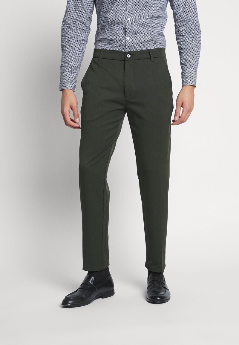 Pantalon vert foncé ajusté en tissu lisse, avec fermeture à bouton et poches latérales, associé à une chemise bleue à motifs et des chaussures noires.