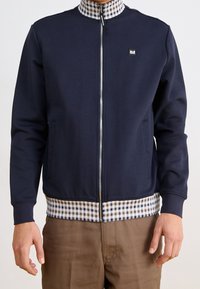 Giacca zip-up blu navy con colletto e orlo a scacchi, dotata di tasche laterali e di un piccolo logo rettangolare sul petto sinistro.