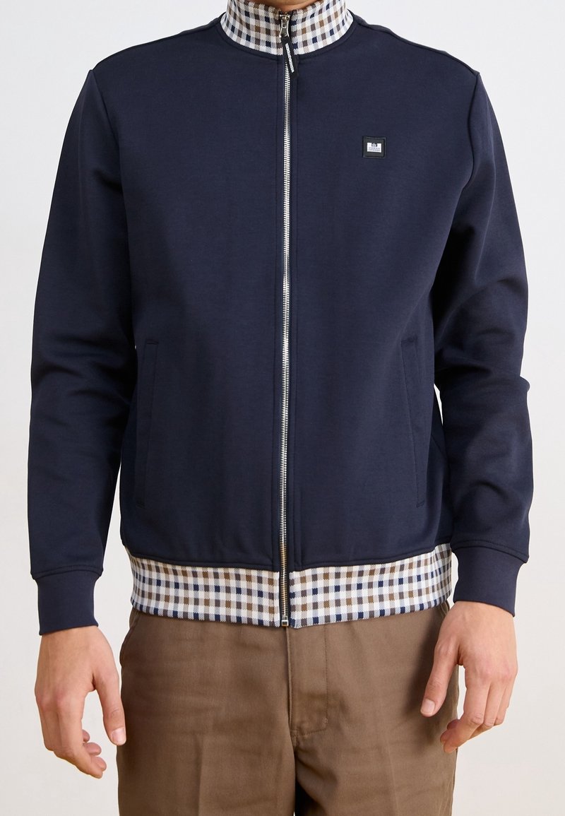 Giacca zip-up blu navy con colletto e orlo a scacchi, dotata di tasche laterali e di un piccolo logo rettangolare sul petto sinistro.
