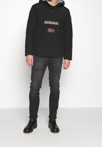 Chubasquero negro con un bolsillo tipo canguro en el frente, parche de logo y una bandera noruega. Combinado con jeans oscuros y botas negras hasta el tobillo.