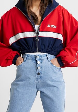 Kvinde iført en kort rød, marineblå og hvid lynlåstrøje og høj talje lyseblå jeans med knaplukning, hænderne i lommerne.