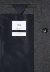 Tessuto grigio testurizzato con foderato in satin nero. Le etichette indicano "MNG", taglia "EUR 50 UK 40" e "SLIM FIT". Un dettaglio con bottone visibile.