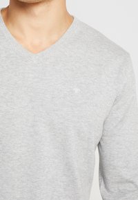 Maglione grigio a V realizzato in tessuto lavorato a maglia, con maniche lunghe e un logo discreto sul petto. Texture morbida con una vestibilità comoda.