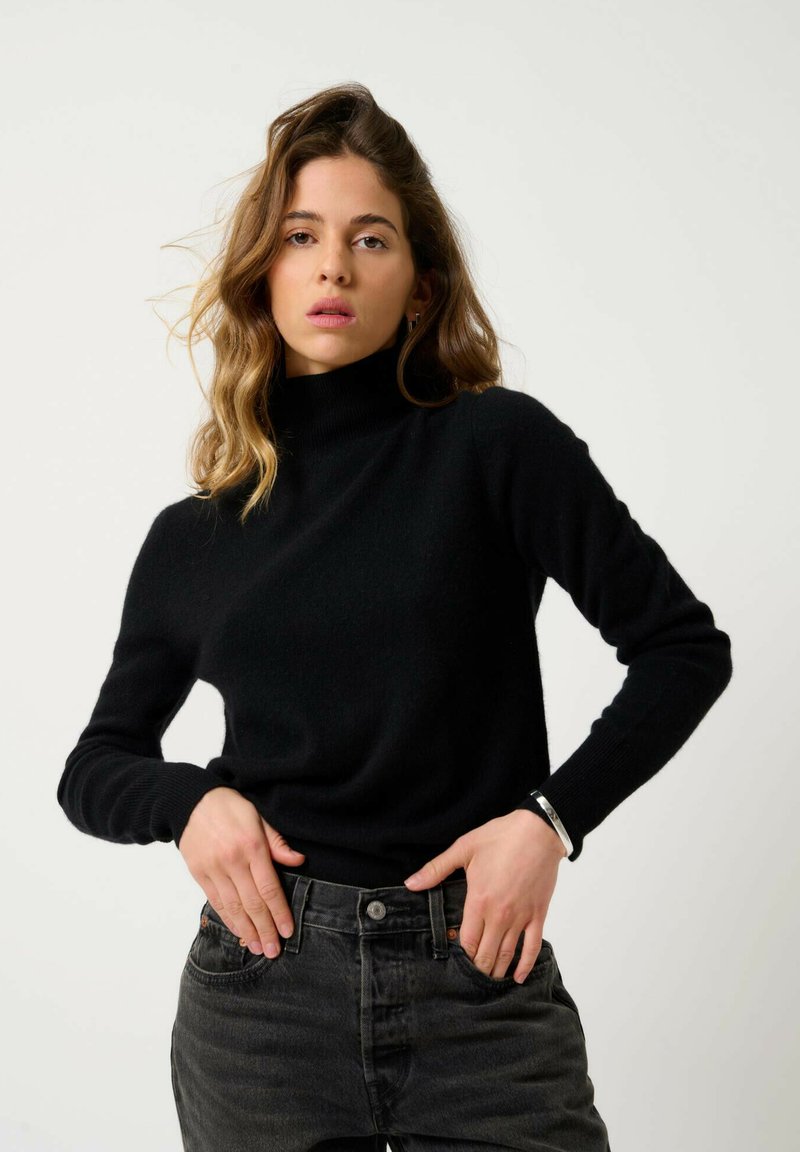 Schwarzer Strickrollkragenpullover mit enganliegenden Ärmeln und weichem Material, kombiniert mit dunkelgrauen Jeans. Keine sichtbaren Muster oder Akzente.