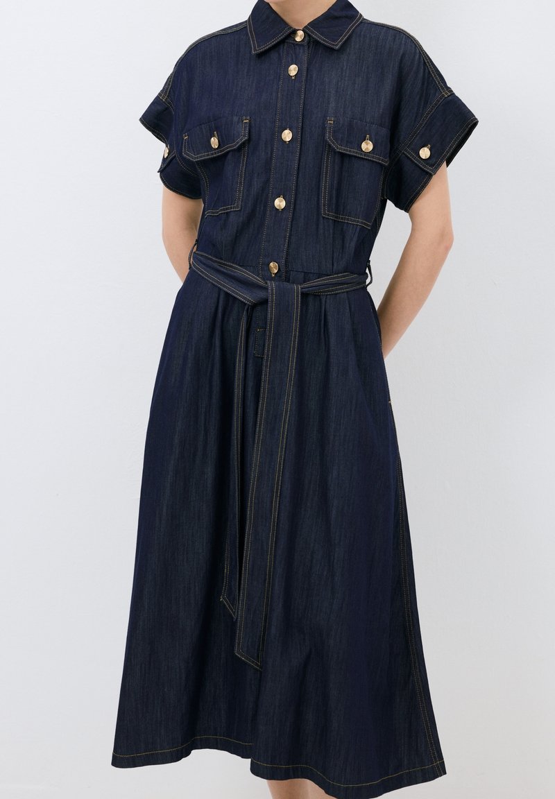 Robe chemise en denim bleu foncé, dotée d'un col, de manches courtes, de deux poches poitrine, de boutons dorés et d'une ceinture assortie.