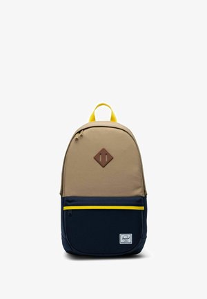 Sac à dos beige et bleu marine avec poignée et fermeture éclair jaunes, poche avant et patch marron en forme de losange sur la partie supérieure.