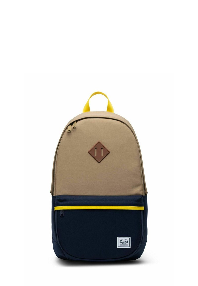 Sac à dos beige et bleu marine avec poignée et fermeture éclair jaunes, poche avant et patch marron en forme de losange sur la partie supérieure.