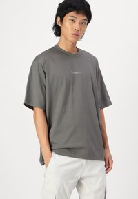 T-shirt en coton gris à coupe décontractée, col rond et manches courtes. Présente des détails de logo subtils sur la poitrine. Design simple.