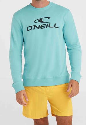 Hombre con sudadera azul claro de O'Neill y pantalones cortos amarillo brillante, de pie frente a un fondo blanco liso.