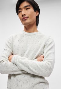 Κρέμαχρωμο πλεκτό πουλόβερ με στίγματα, ριγωτές μανσέτες και στρογγυλή λαιμόκοψη, φορεμένο από μοντέλο με σταυρωμένα χέρια.