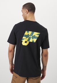 Black cotton t-shirt featuring a large geometric graphic in green and yellow on the back, with a rectangular pattern and bold shapes.