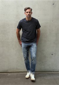 Mörkgrå kortärmad skjorta, ljusblå jeans och vita sportskor med beige detaljer, mot en betongvägg i bakgrunden.