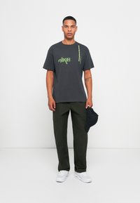 Mørkegrå Nike t-shirt med grønt logo, parret med olivengrønne cargobukser og hvide sneakers, mens han/hun holder en sort kasket.