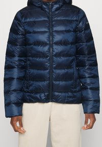 Man med en marinblå vadderad jacka med dragkedja fram och beige byxor, stående mot en ljus bakgrund.