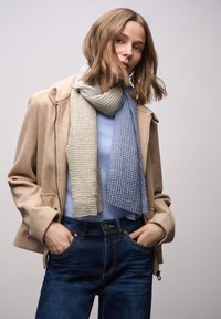 Tørklæde i lysegrøn og blå med et tekstureret, stribet mønster. Båret over en blå top, kombineret med en beige jakke og mørke jeans.