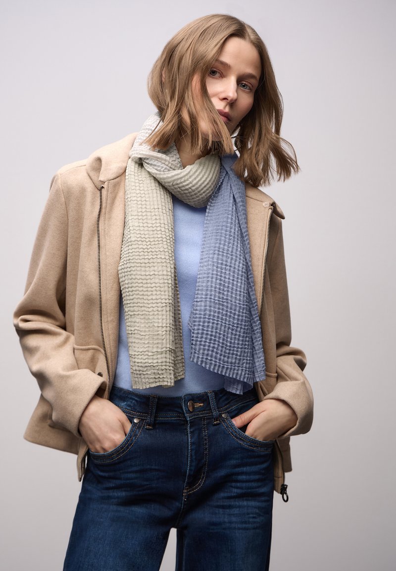 Tørklæde i lysegrøn og blå med et tekstureret, stribet mønster. Båret over en blå top, kombineret med en beige jakke og mørke jeans.