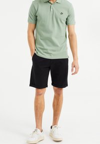 Polo en vert pâle avec un col et un logo ; shorts noirs avec les ourlets retroussés ; baskets blanches avec des accents gris clair.