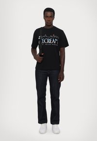Black cotton t-shirt with "Scream" graphic, paired with dark jeans and white sneakers. Model holds a black strap bag.