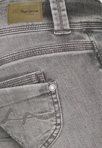 Tascone dei jeans in denim grigio con cuciture visibili, rivetto metallico e un'etichetta in pelle marrone chiaro con la scritta "Pepe Jeans London" all'altezza della vita.